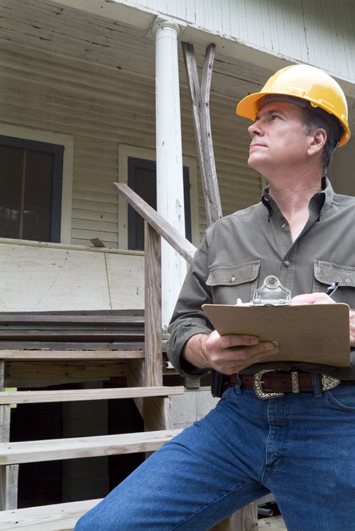 A man in a hard hat standing in front of an old rundown house holding a clipboard in his hand.